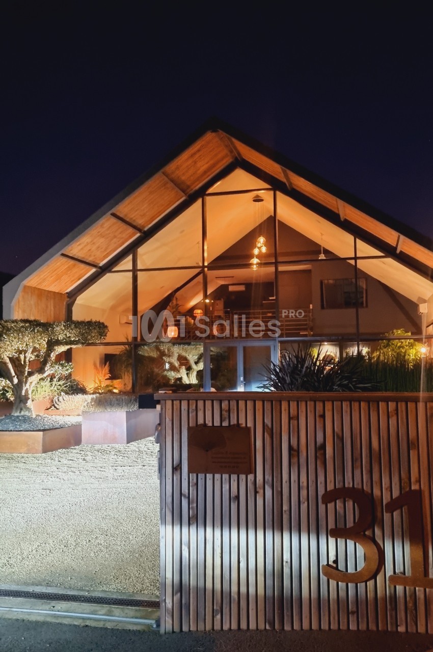 Façade d'une maison moderne en bois et verre, éclairée de nuit, avec jardin et portail en premier plan.