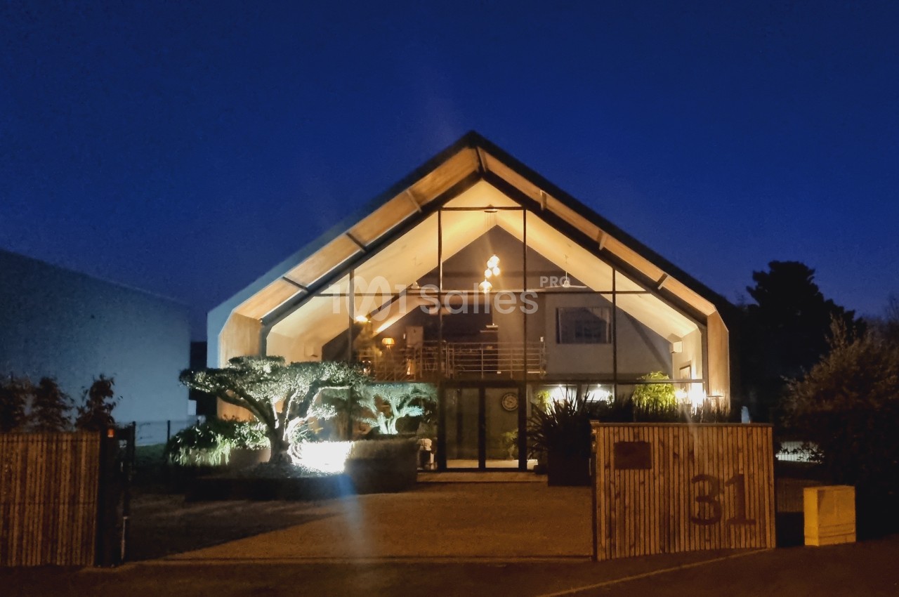 Façade d'un bâtiment moderne avec une grande verrière illuminée, entouré de végétation, photographié de nuit.