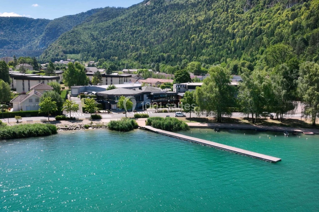 Vue aérienne d'un lac turquoise bordé de végétation, avec un ponton et des bâtiments en arrière-plan.