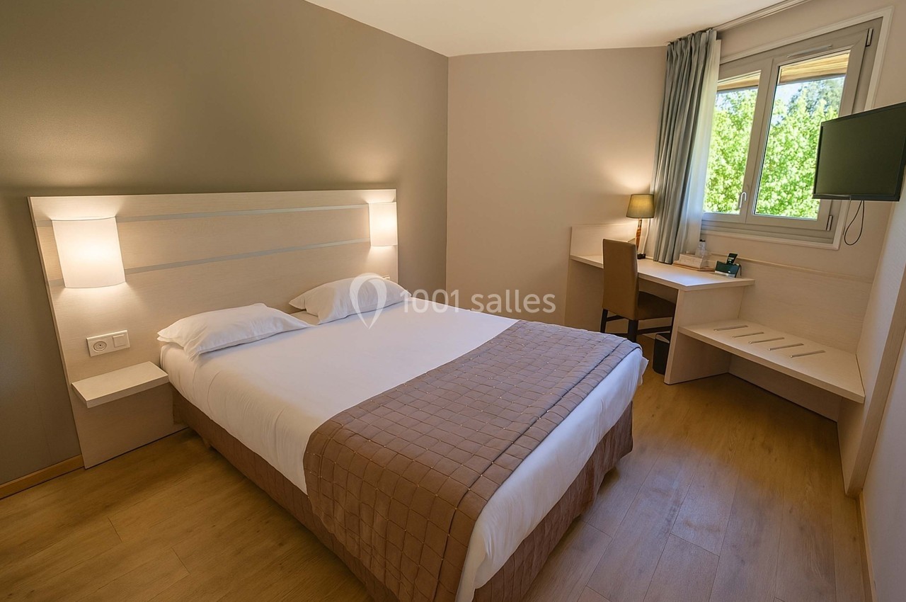 Chambre lumineuse avec lit double, bureau, télévision murale et fenêtre donnant sur un espace verdoyant.
