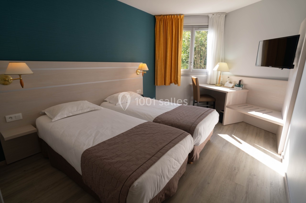 Chambre lumineuse avec deux lits simples, bureau, télévision murale, rideaux jaunes et fenêtre donnant sur l'extérieur.