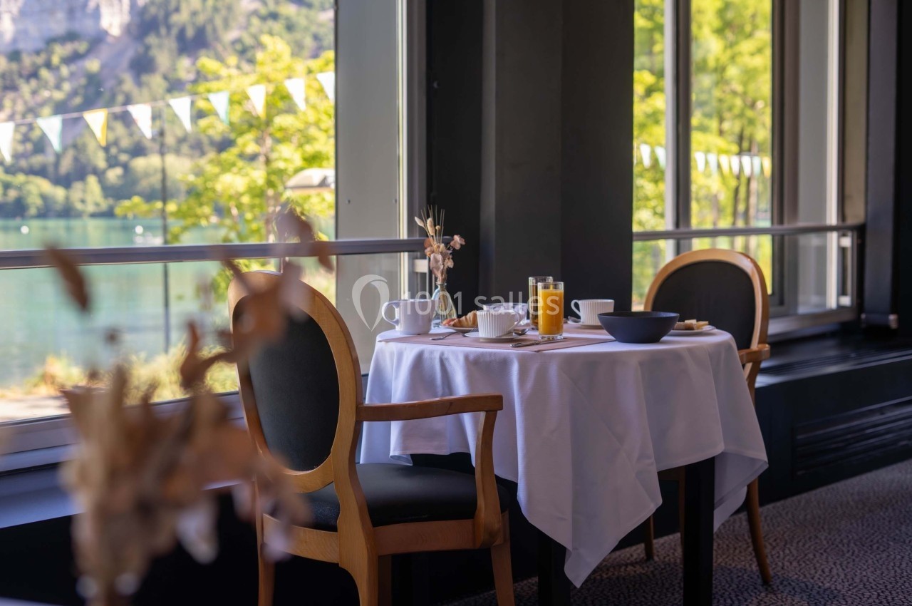 Table dressée avec petit-déjeuner, vue sur un lac et des arbres à travers de grandes fenêtres lumineuses.