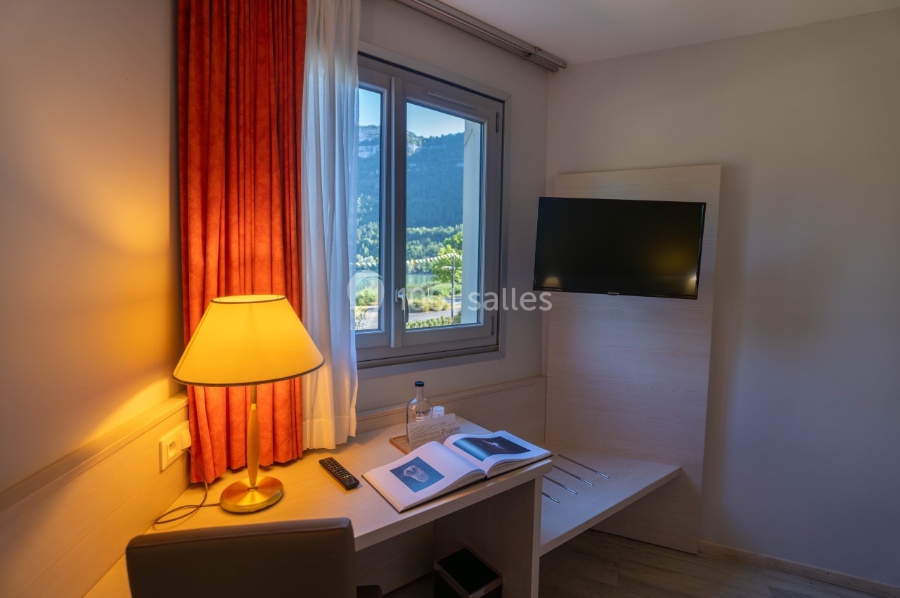 Bureau d'hôtel avec lampe allumée, livre ouvert, télévision murale et fenêtre offrant une vue sur un paysage naturel.
