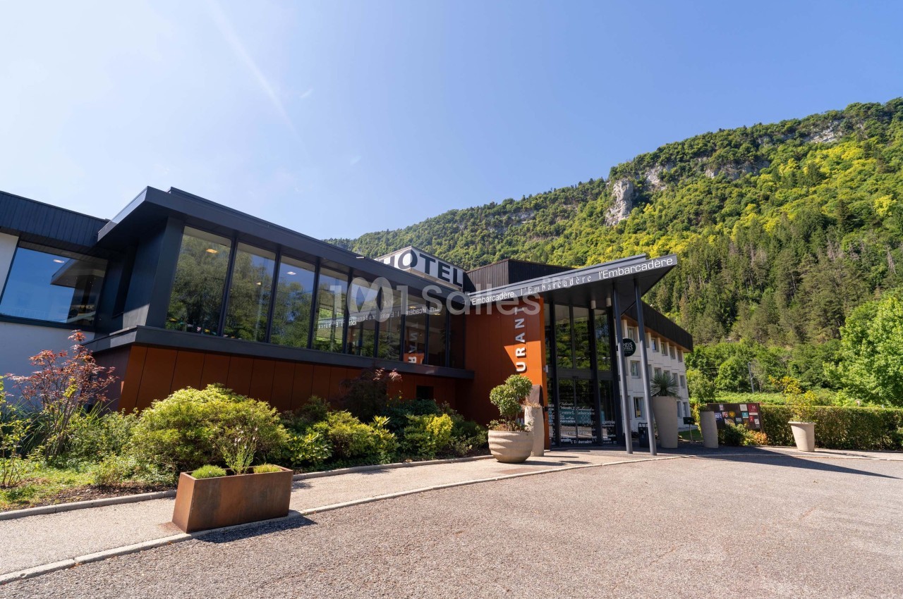 Façade moderne d'un hôtel entouré de végétation, avec une colline boisée en arrière-plan sous un ciel dégagé.