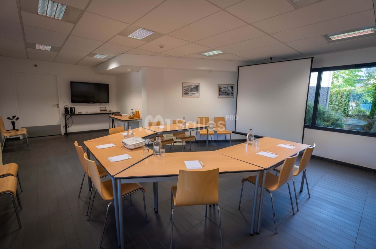 Salle de réunion lumineuse avec tables disposées en U, chaises en bois, écran de projection et matériel de présentation.