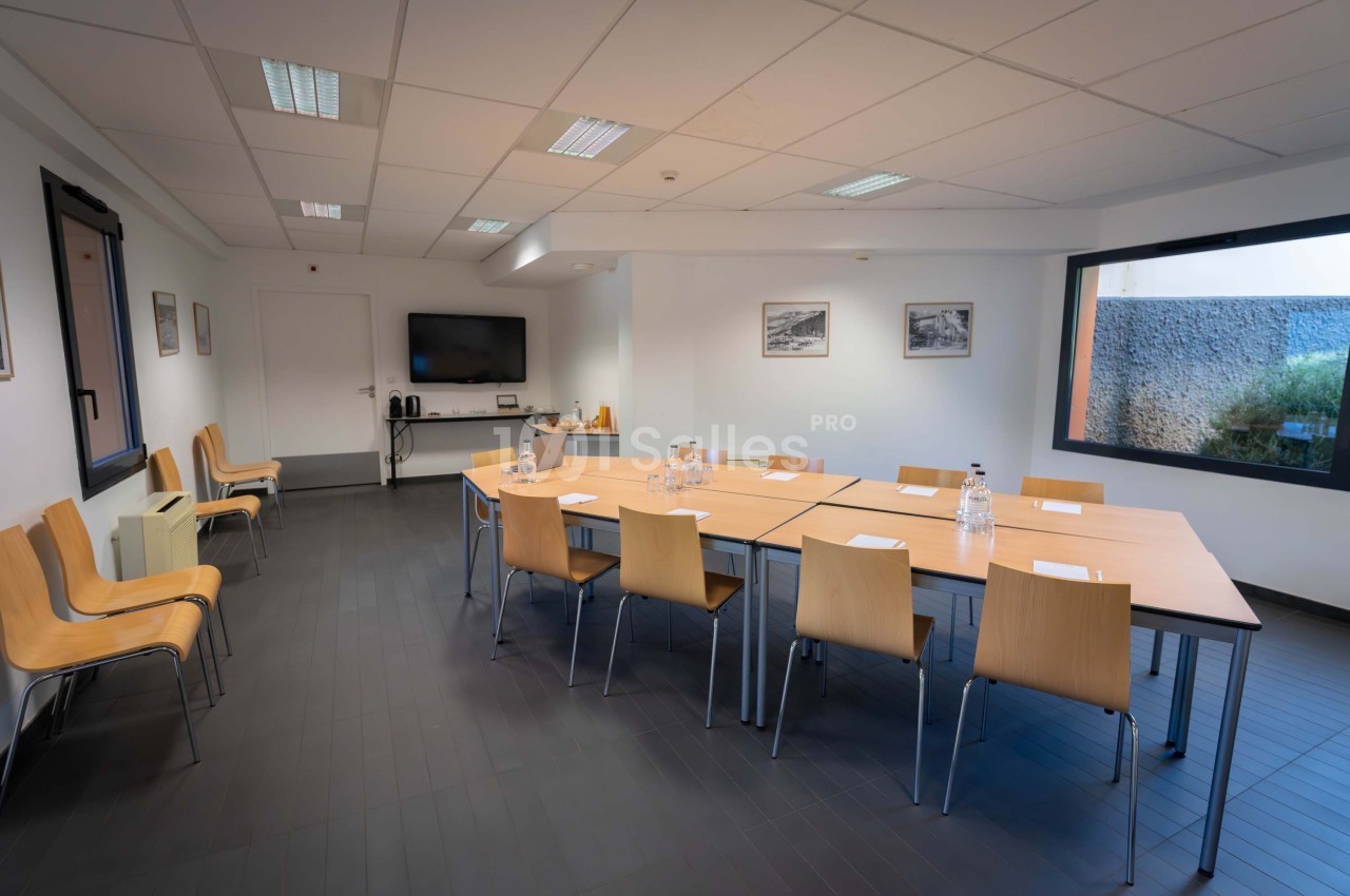 Salle de réunion lumineuse avec table rectangulaire, chaises en bois, écran mural et fenêtres donnant sur l'extérieur.