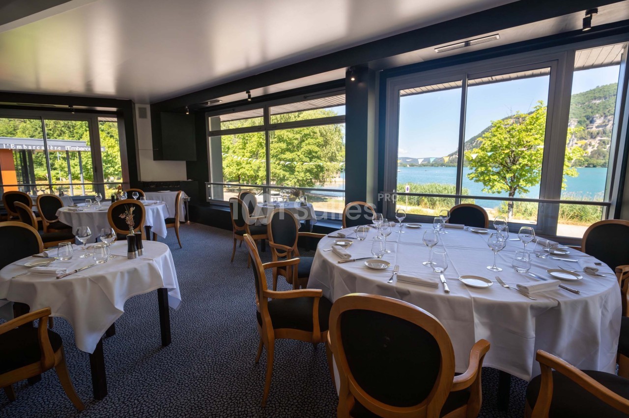 Salle de restaurant lumineuse avec tables dressées, grandes baies vitrées offrant une vue sur un lac et des collines…