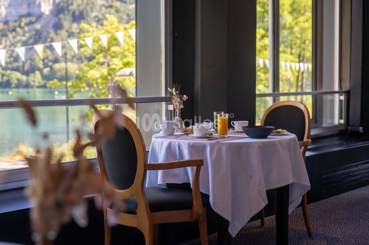 Table dressée avec petit-déjeuner, vue sur un lac et des arbres à travers de grandes fenêtres lumineuses.