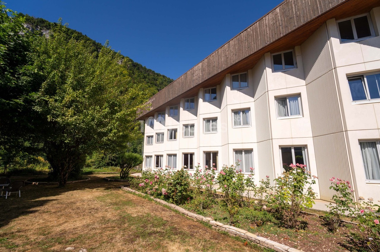 Façade d'un bâtiment moderne entouré de rosiers et d'arbres, avec un ciel dégagé en arrière-plan.