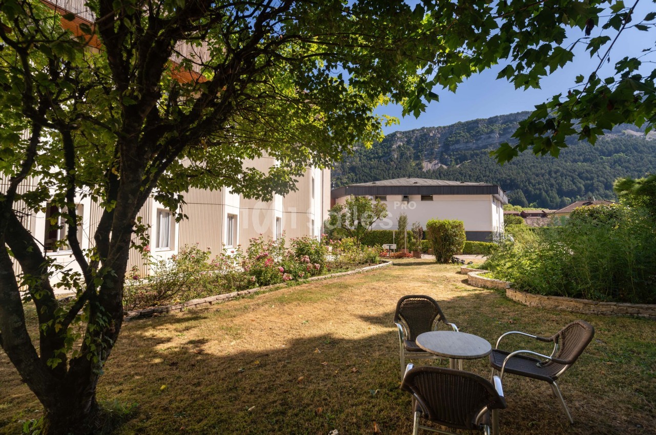 Jardin avec pelouse, arbres, table et chaises, entouré de bâtiments et montagnes en arrière-plan.