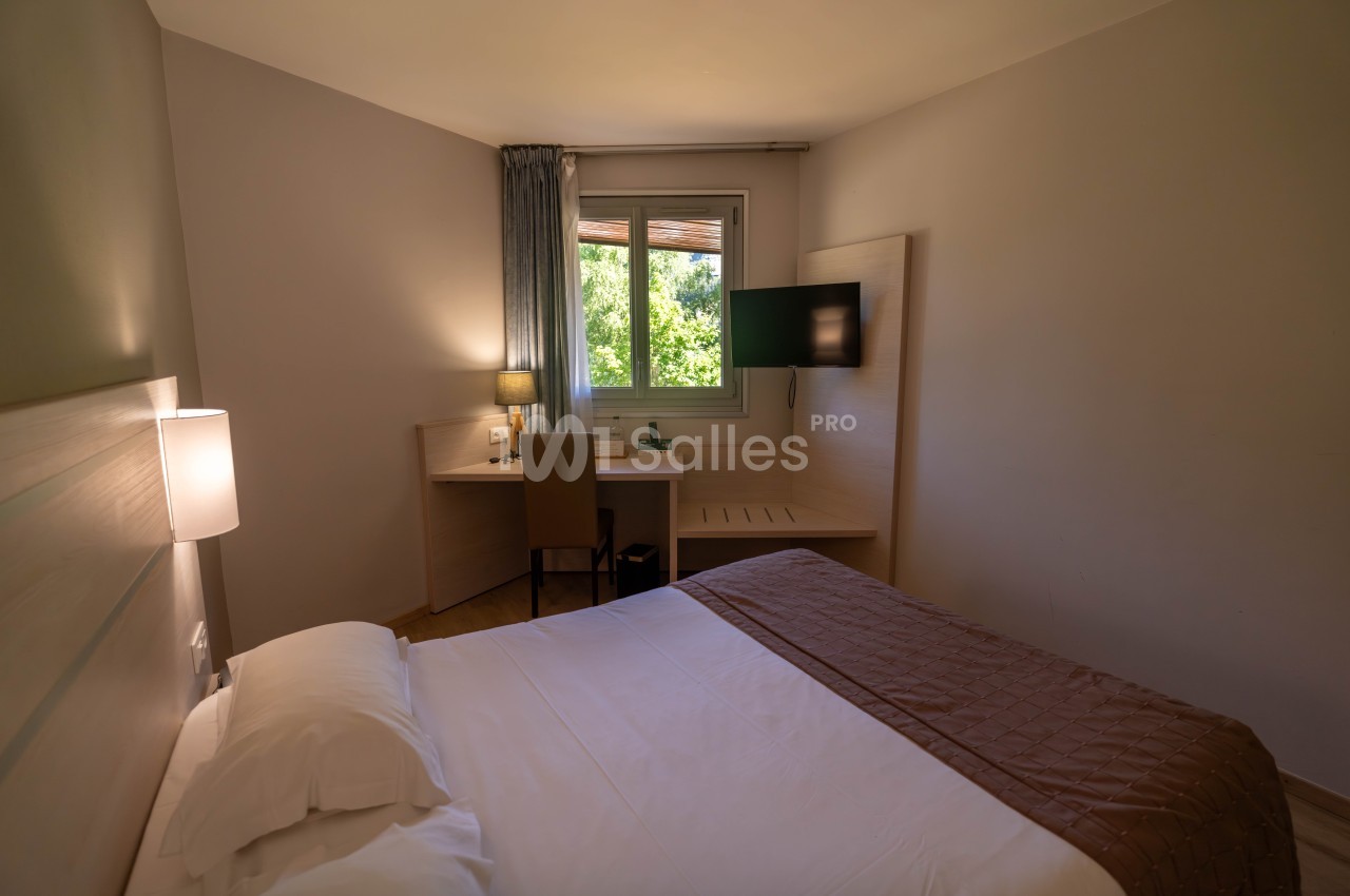 Chambre lumineuse avec lit double, bureau, chaise, télévision murale et fenêtre donnant sur un espace vert.