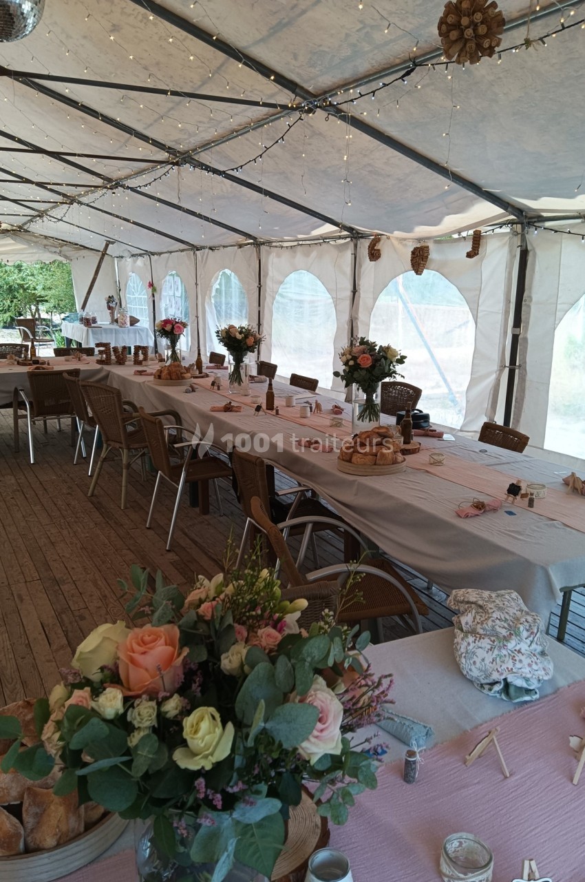 Salle de réception sous chapiteau avec tables décorées de nappes, fleurs et guirlandes lumineuses suspendues.