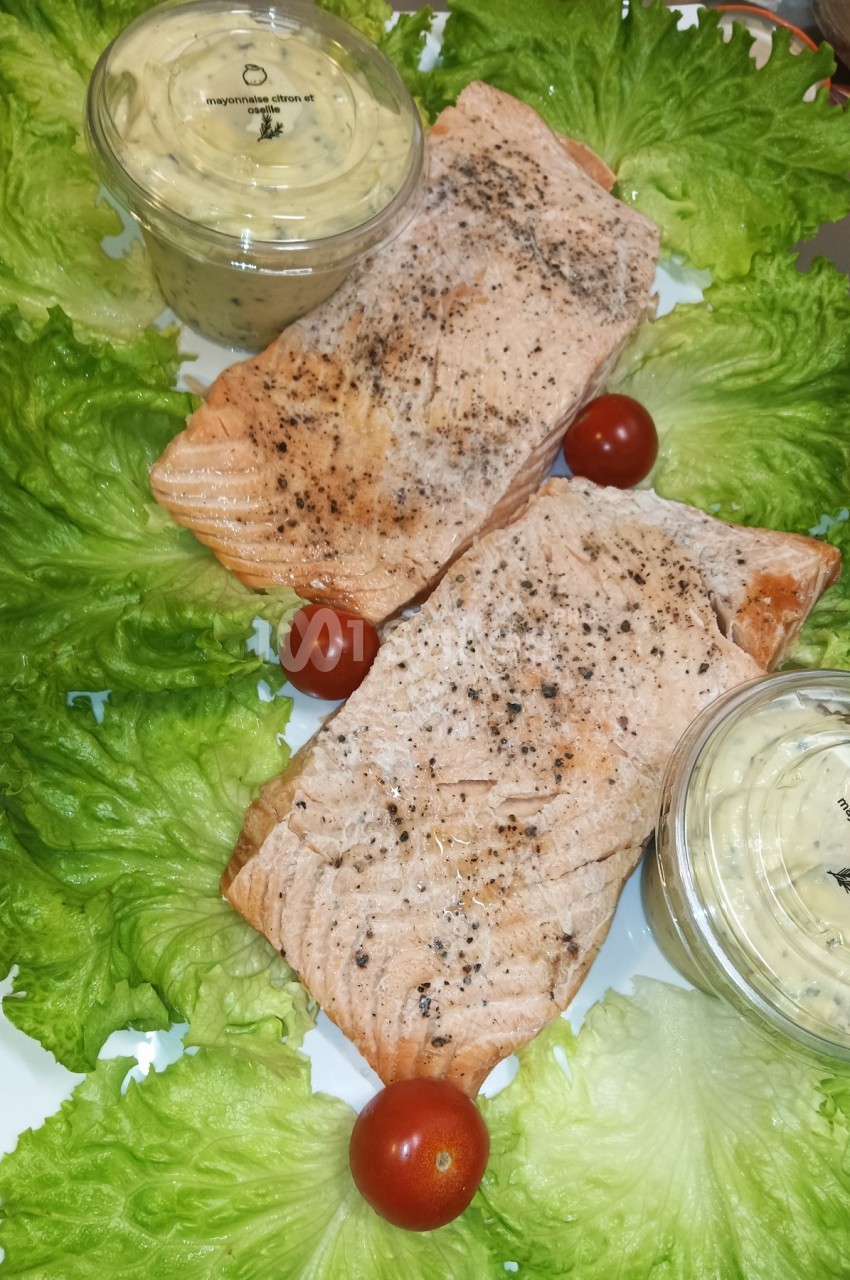 Deux filets de saumon cuits, assaisonnés, disposés sur un lit de laitue avec des tomates cerises et des sauces en pot.