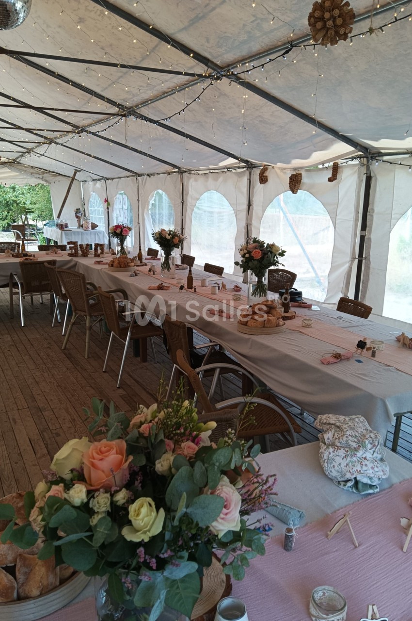Salle de réception sous chapiteau avec tables décorées de nappes, bouquets de fleurs et guirlandes lumineuses.