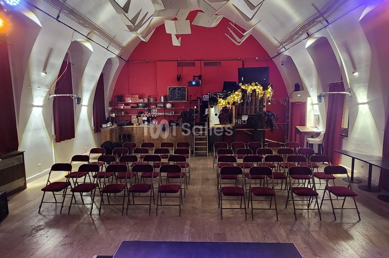 Salle de spectacle avec rangées de chaises rouges, scène au premier plan et bar en fond dans un décor rouge et blanc.