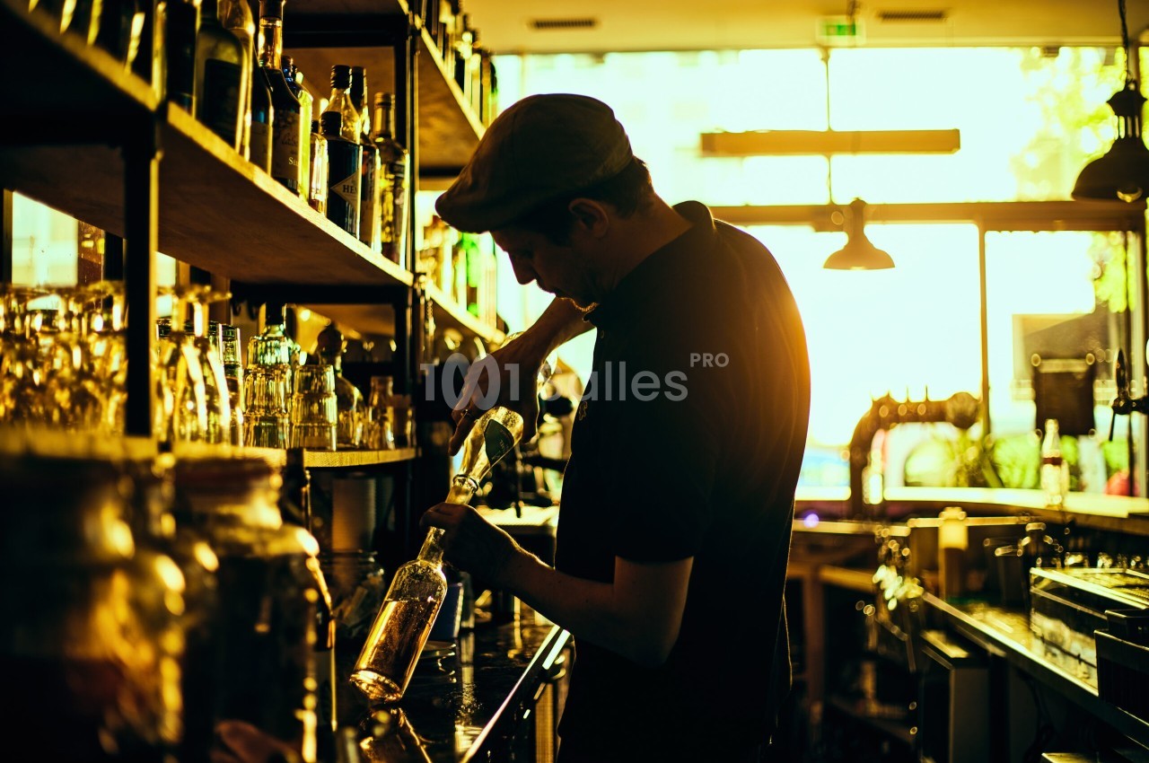Un barman verse une boisson dans une bouteille derrière un comptoir, éclairé par une lumière dorée en fin de journée.