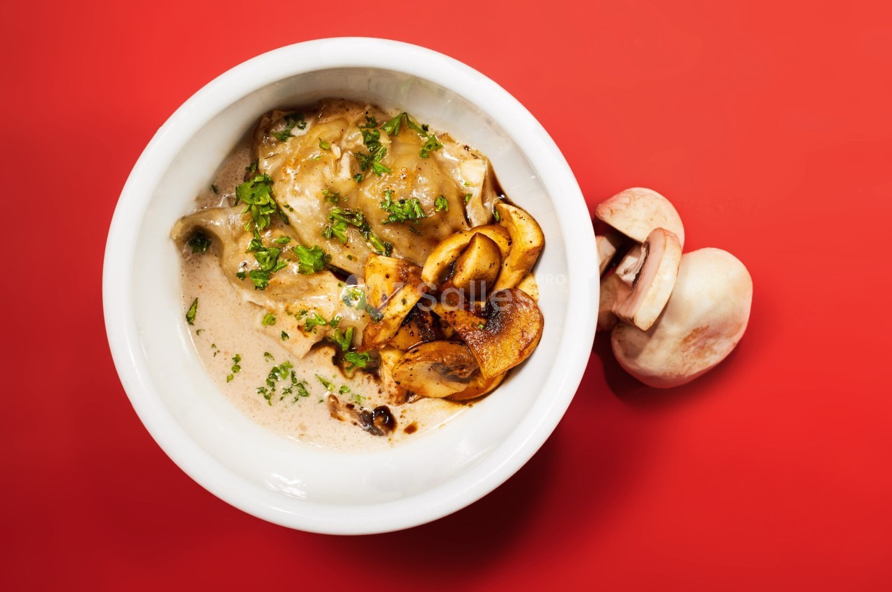 Raviolis dans une sauce crémeuse, garnis de champignons poêlés et de persil, sur fond rouge.