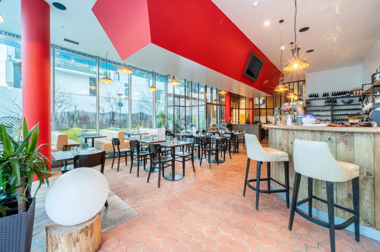 Salle de restaurant lumineuse avec grandes baies vitrées, mobilier moderne, bar en bois et plafond rouge.