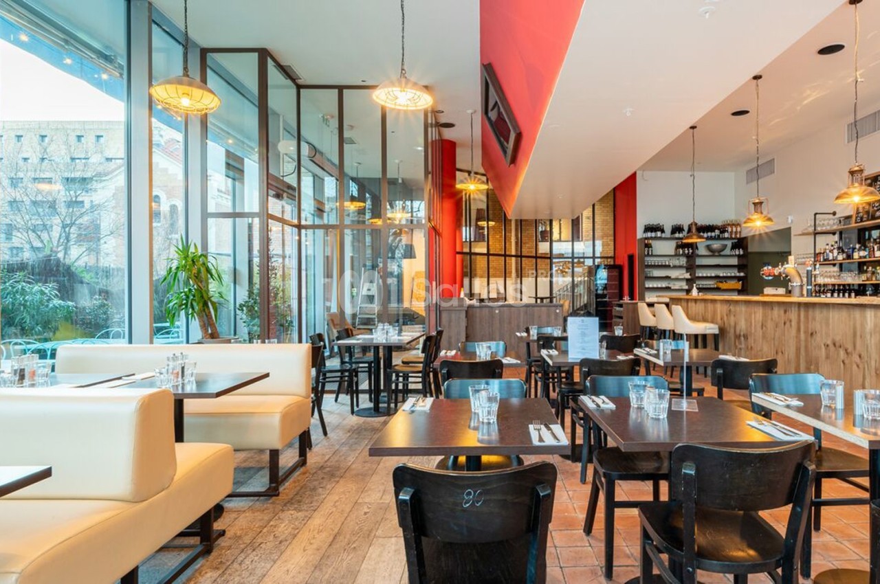 Salle de restaurant lumineuse avec grandes baies vitrées, mobilier en bois foncé et banquettes claires.