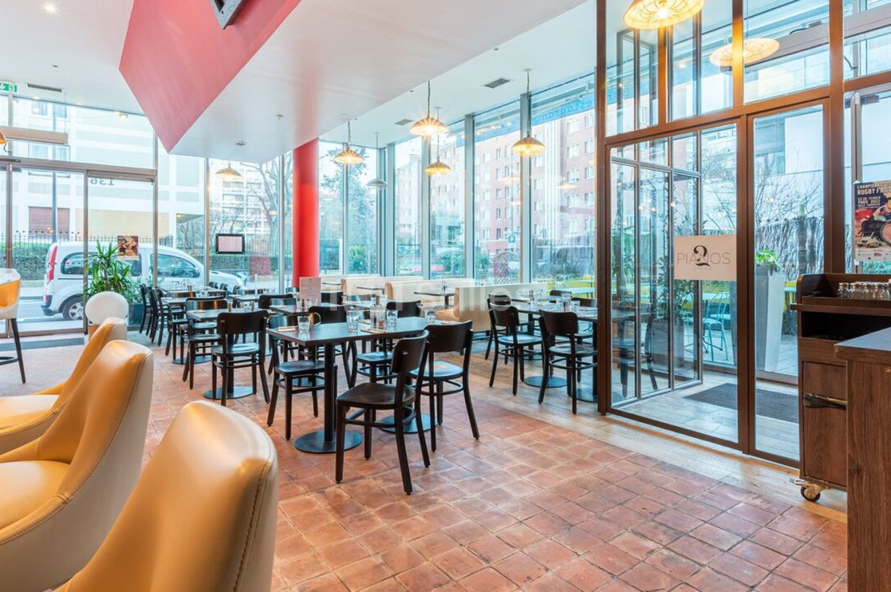 Salle de restaurant lumineuse avec tables et chaises noires, grandes baies vitrées et décor moderne.