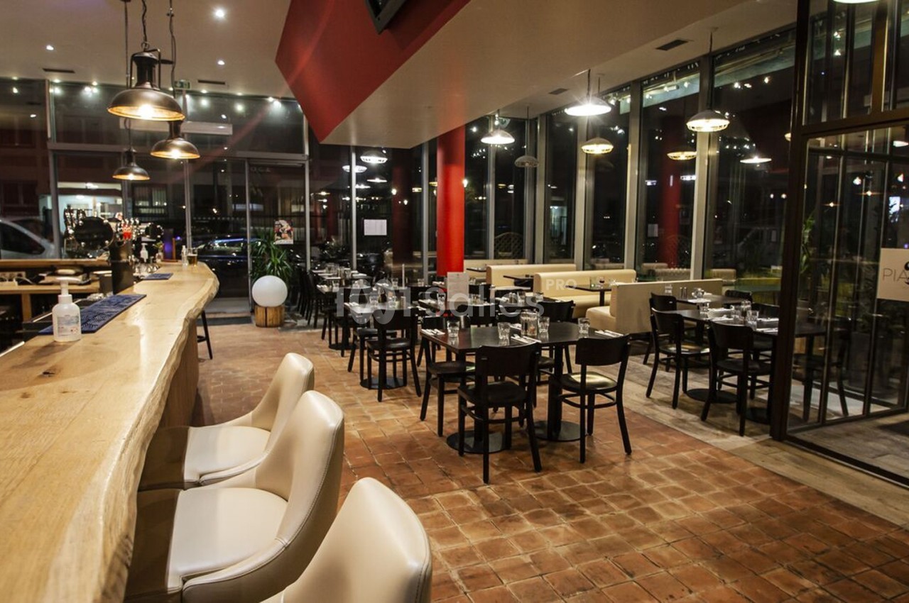 Salle de restaurant moderne avec bar en bois, tables dressées et grandes baies vitrées donnant sur l'extérieur.