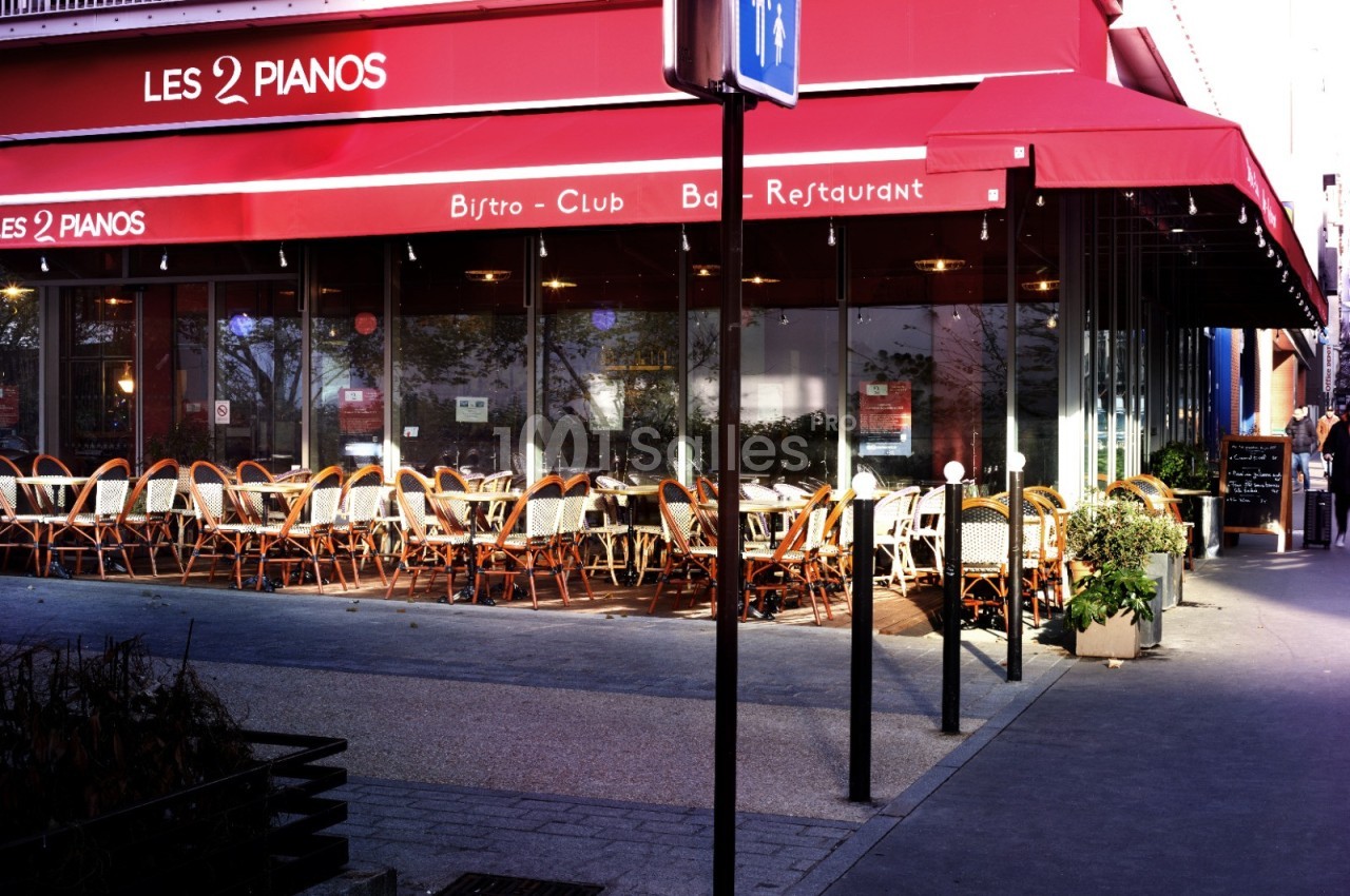 Terrasse d'un restaurant avec des chaises en osier et une devanture rouge, partiellement ombragée par un auvent.