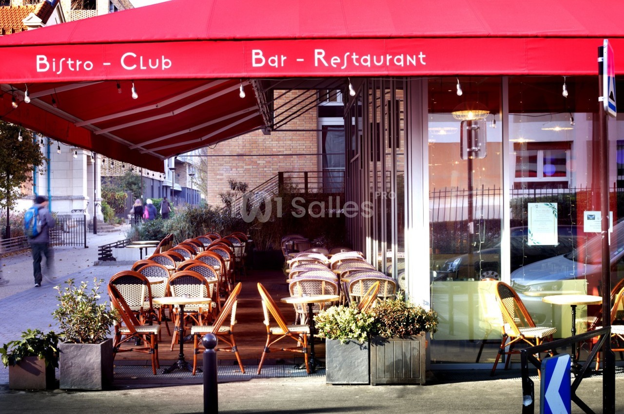 Terrasse d'un bistro avec des chaises en osier sous un auvent rouge, située près d'une rue piétonne.