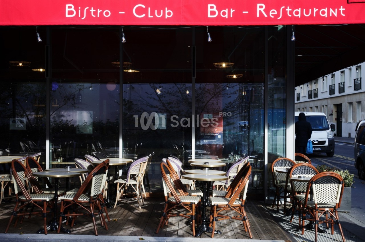 Terrasse extérieure d'un bistro avec des tables et chaises en osier sous un auvent rouge, vue sur la rue adjacente.