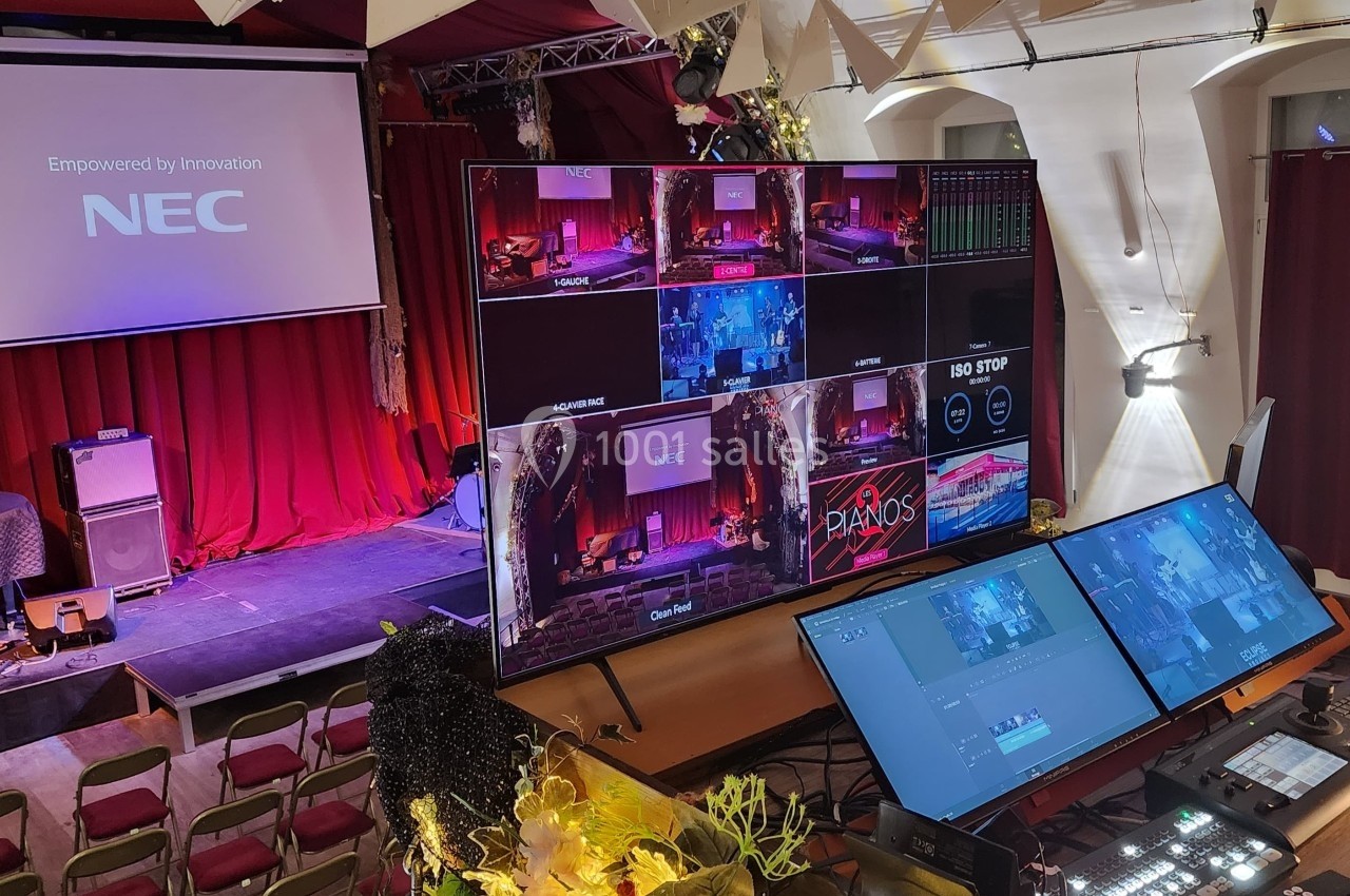 Vue d'une régie audiovisuelle avec plusieurs écrans et un plateau de scène équipé, dans une salle de spectacle.