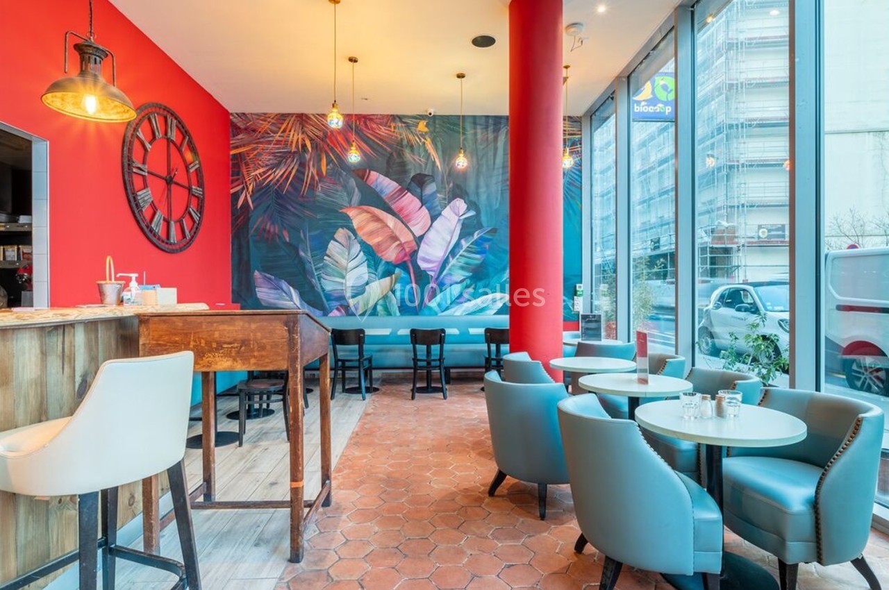 Salle de restaurant lumineuse avec murs rouges, décor tropical, mobilier moderne et grandes baies vitrées.