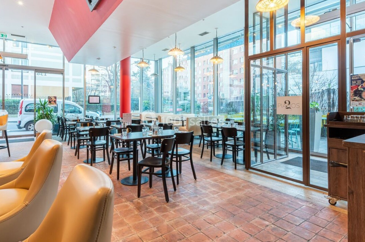 Salle de restaurant lumineuse avec tables et chaises noires, grandes baies vitrées et décor moderne.