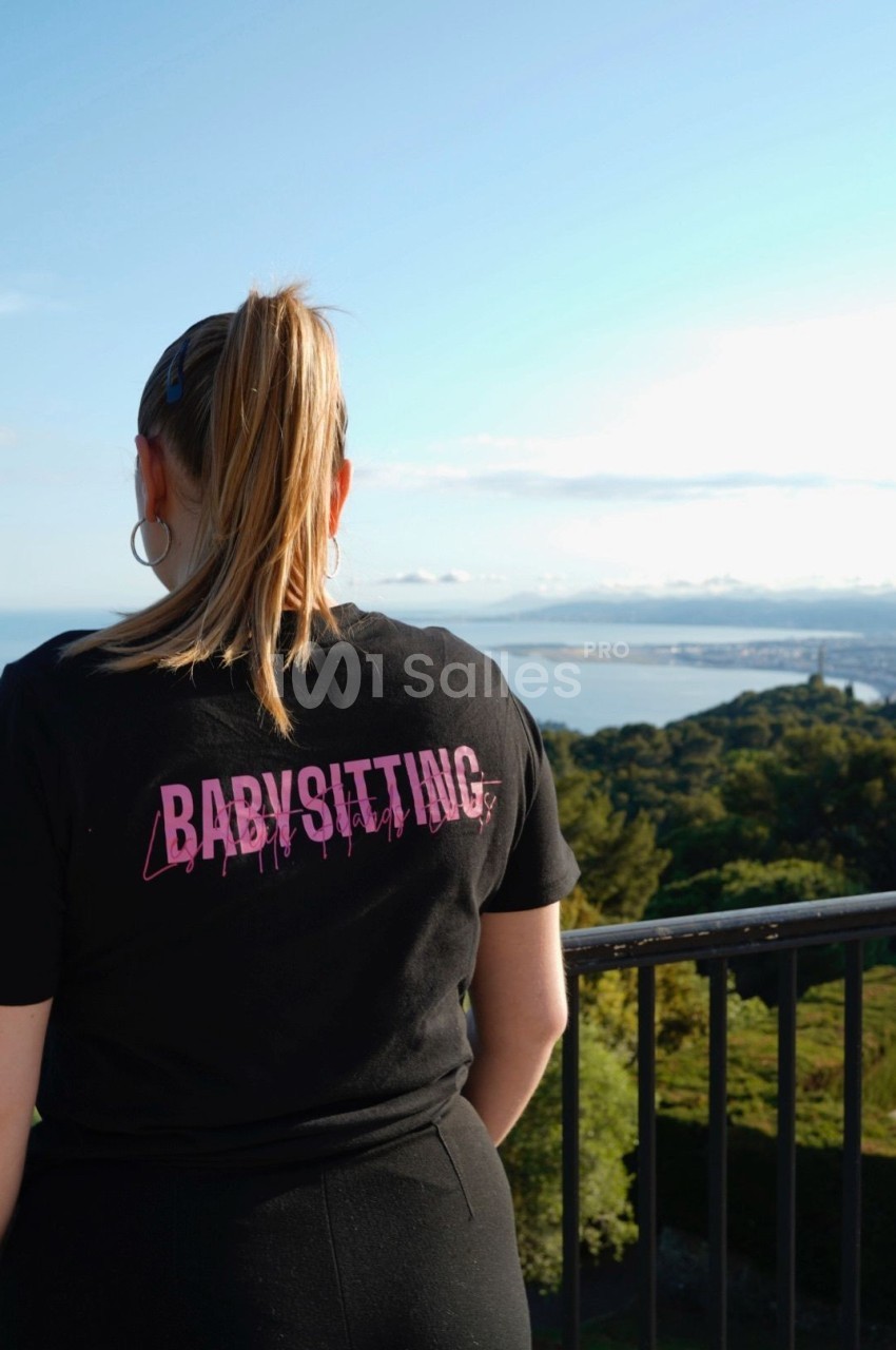 Une femme de dos, portant un t-shirt noir avec l'inscription ’Babysitting’, devant un paysage côtier.