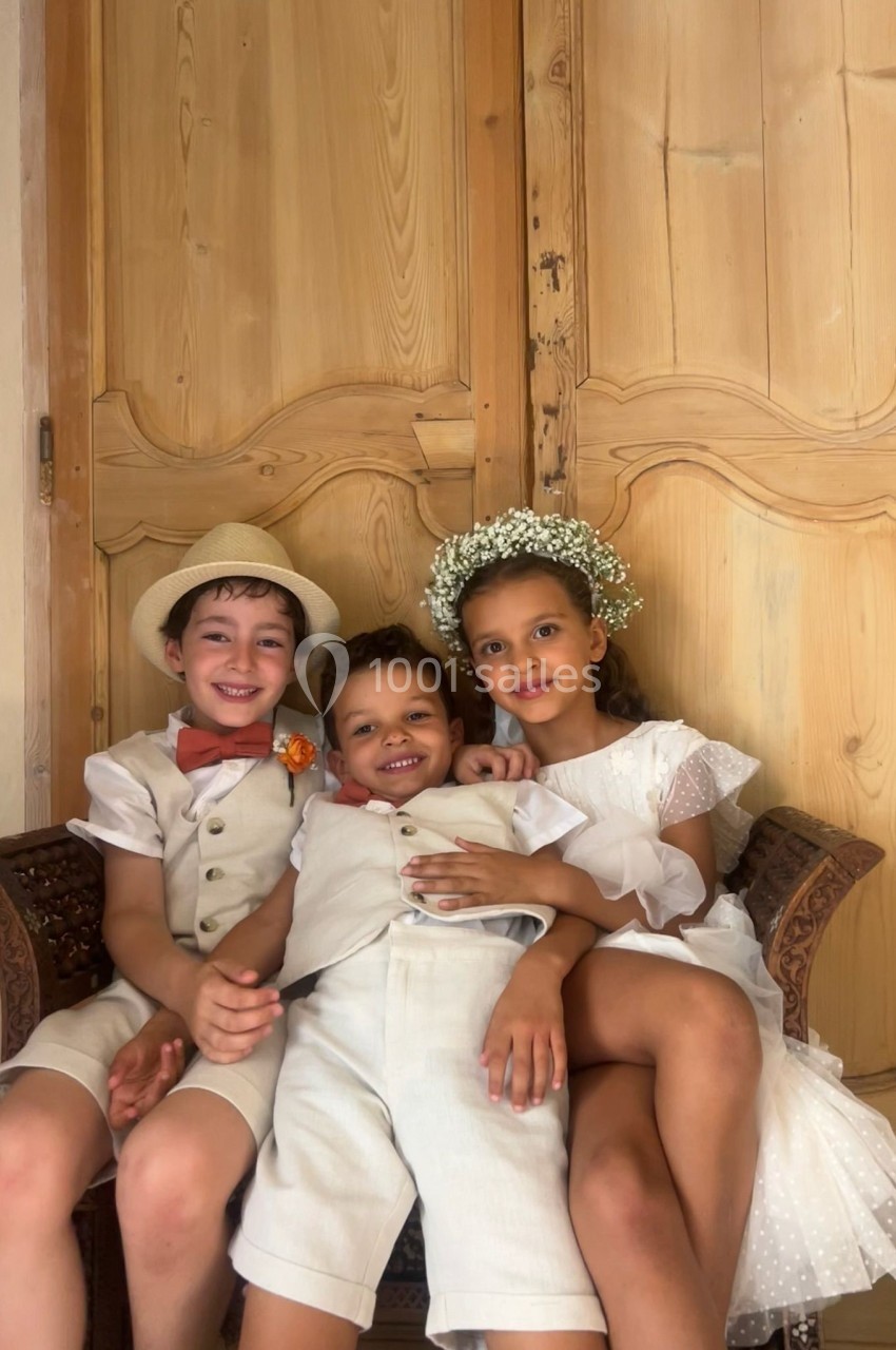 Trois enfants souriants en tenue élégante, assis sur un fauteuil devant une porte en bois sculpté.
