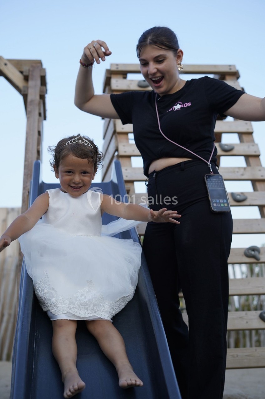 Une petite fille en robe blanche glisse sur un toboggan, accompagnée d'une femme souriante en arrière-plan.