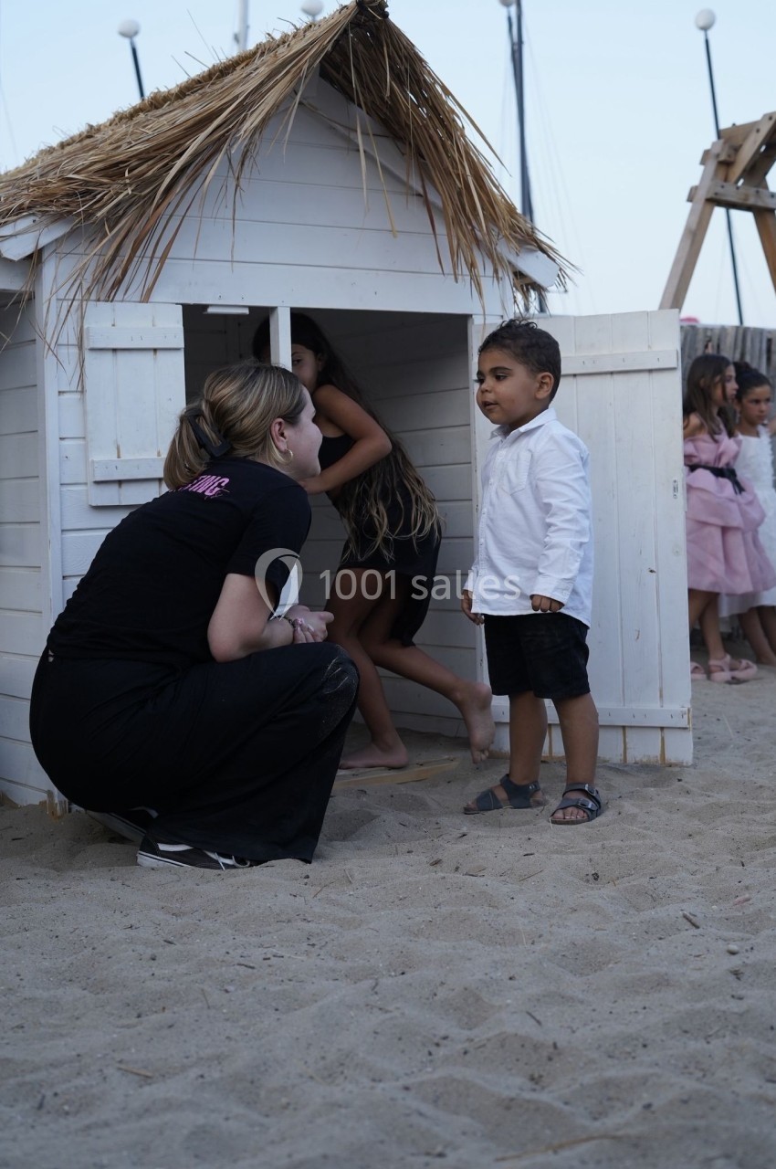 Une femme accroupie parle à un jeune garçon devant une cabane en bois sur une plage, avec des enfants en arrière-plan.