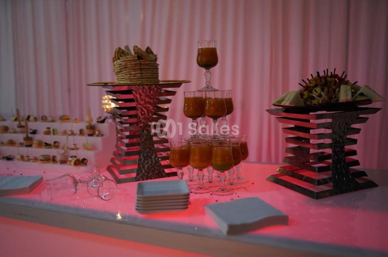Buffet avec verrines de soupe, brochettes de fruits, biscuits et serviettes sur une table éclairée en rose.
