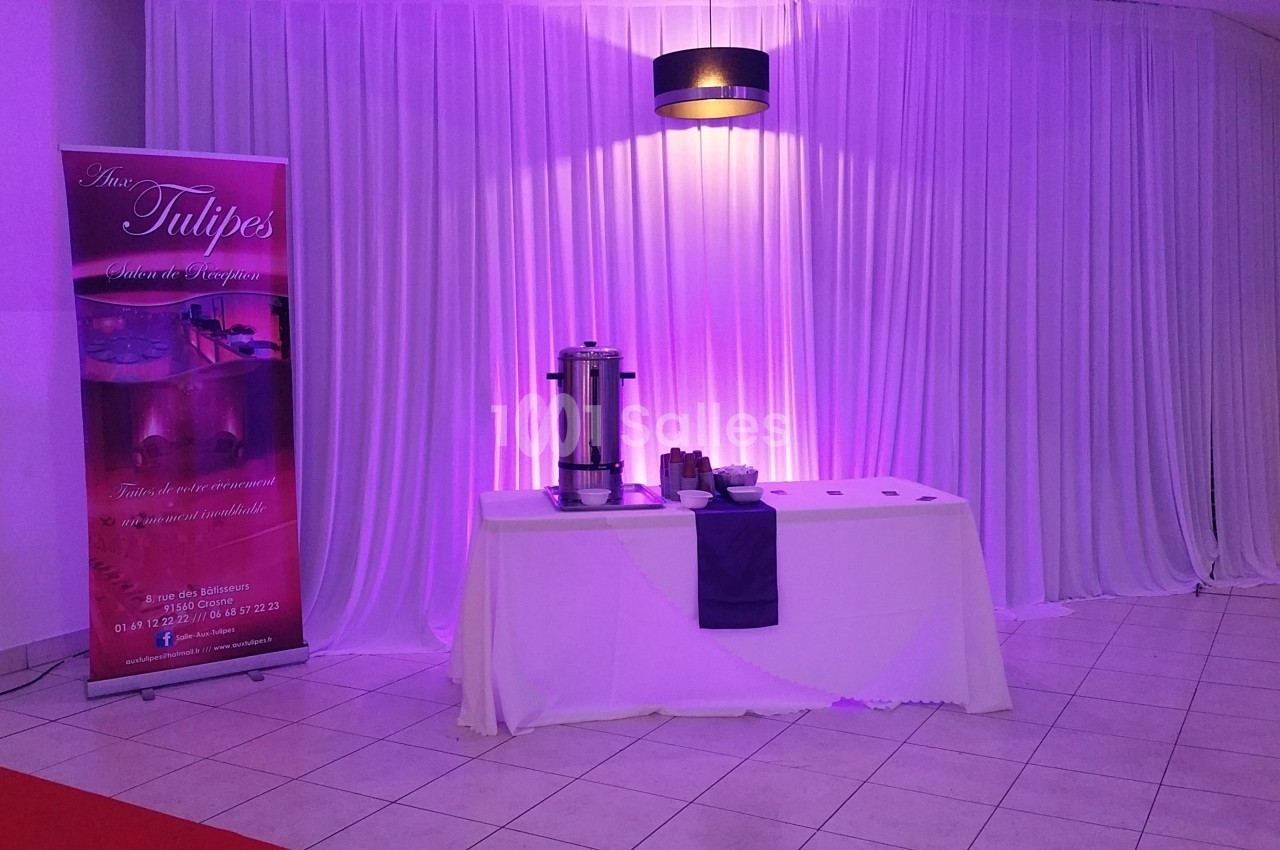 Table dressée avec une nappe blanche, urne à café et accessoires, devant un rideau blanc éclairé de lumière violette.