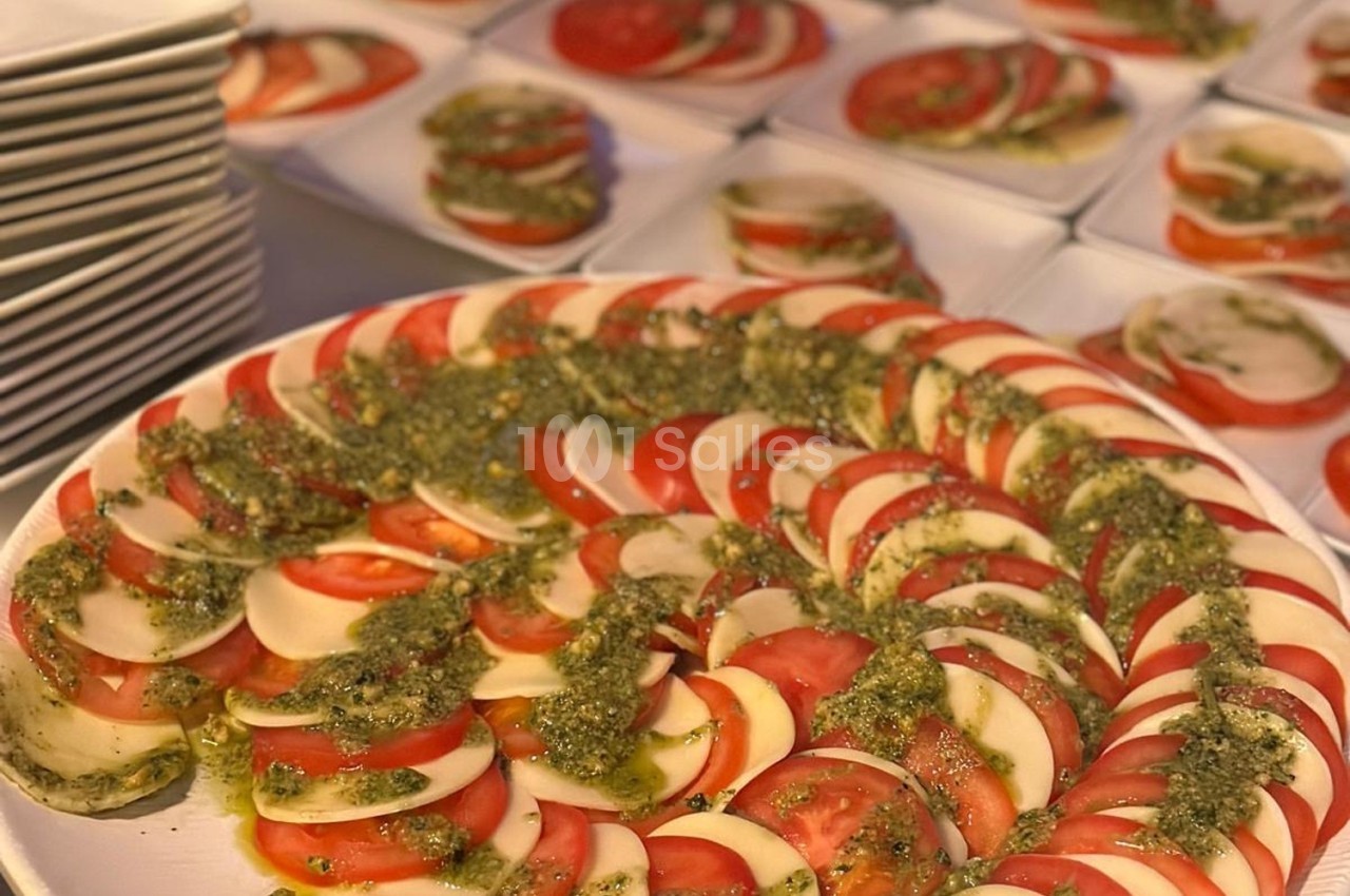 Assiette de salade caprese avec des tranches de tomate, mozzarella et pesto, entourée d'autres assiettes similaires.