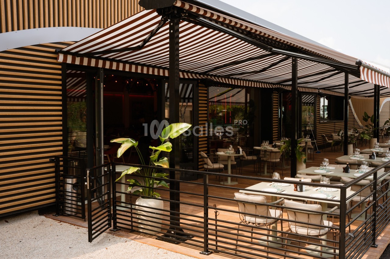 Terrasse d'un restaurant avec des tables dressées, des plantes en pot et des stores rayés noirs et blancs.