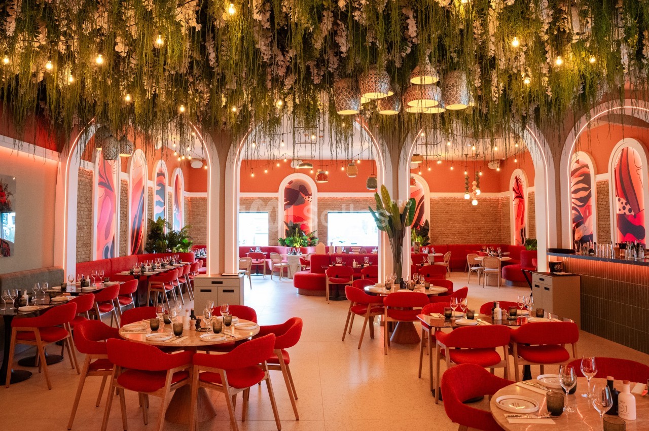 Salle de restaurant lumineuse avec des tables dressées, des chaises rouges et un plafond décoré de plantes suspendues.