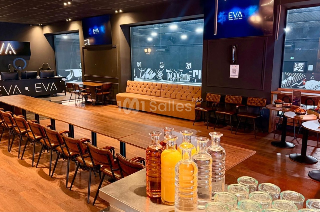 Salle spacieuse avec une longue table en bois, chaises, canapés, écrans et un comptoir avec boissons et carafes.