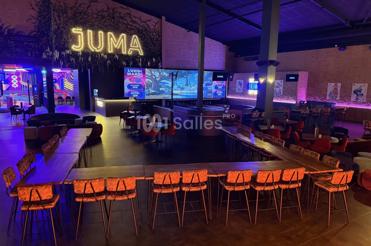 Salle moderne avec tables, chaises et bar central, éclairée par des néons et un grand écran en arrière-plan.