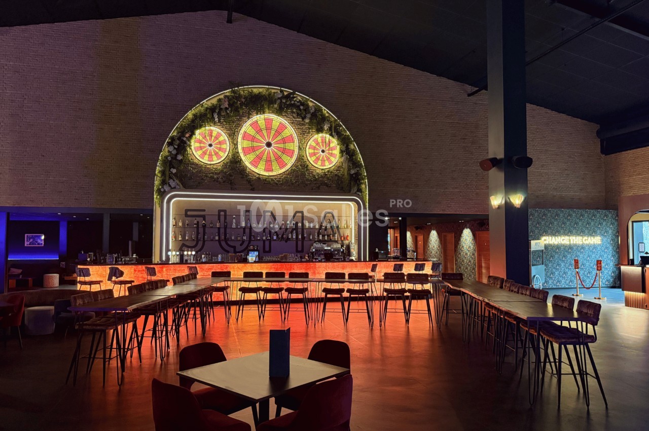 Intérieur d'un bar avec un long comptoir éclairé, des tables et des chaises, et une décoration lumineuse au mur.