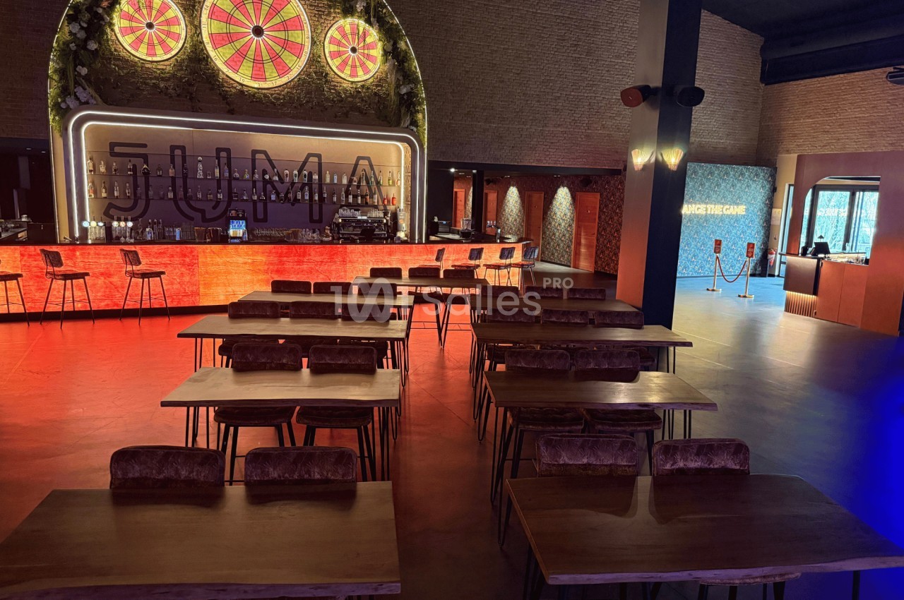 Salle avec tables en bois, chaises rembourrées, bar éclairé en arrière-plan et décorations lumineuses.