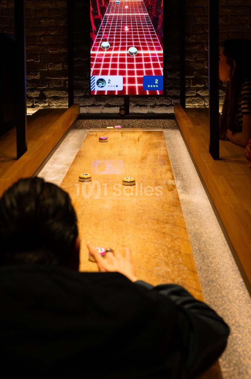 Personnes jouant à un jeu interactif de palets sur une table en bois, avec un écran affichant le score en arrière-plan.