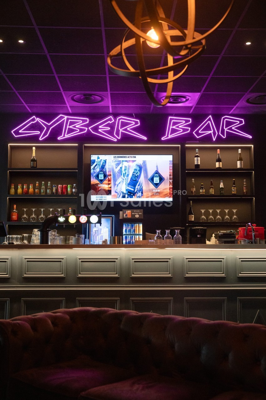 Bar moderne avec néons violets ’Cyber Bar’, comptoir éclairé, étagères de boissons et écrans affichant des publicités.