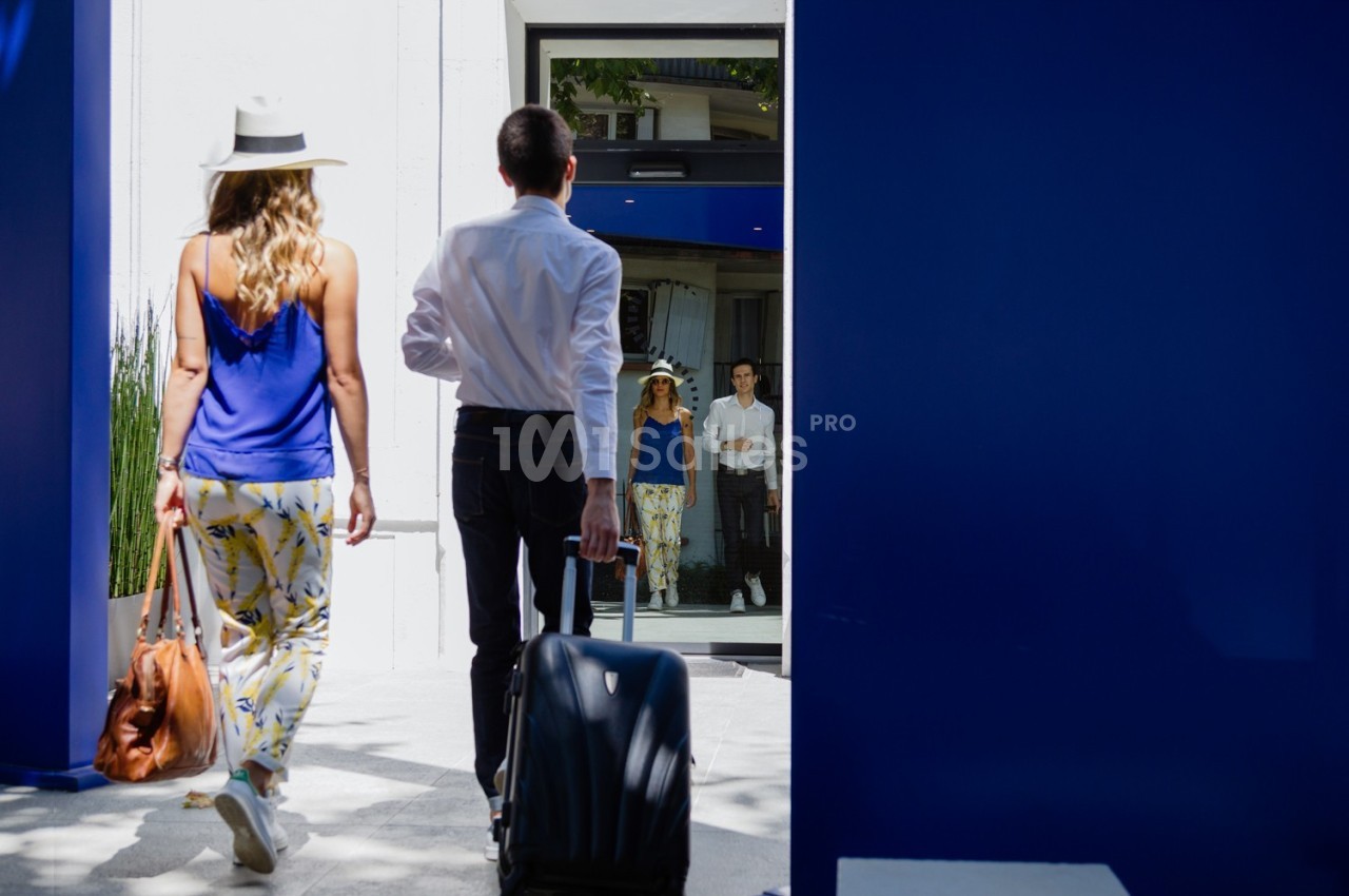 Un homme et une femme entrent dans un bâtiment moderne, lui portant une valise, elle un sac à main.