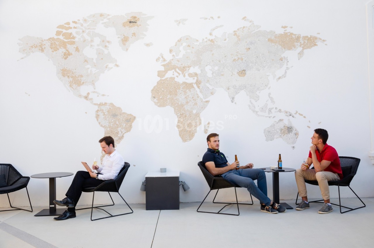 Trois hommes assis sur des chaises devant une carte du monde murale, discutant ou utilisant un ordinateur portable.