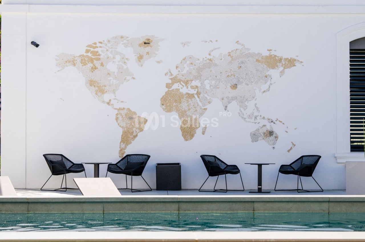 Mur blanc avec une carte du monde texturée, entouré de chaises noires et de petites tables près d'une piscine.