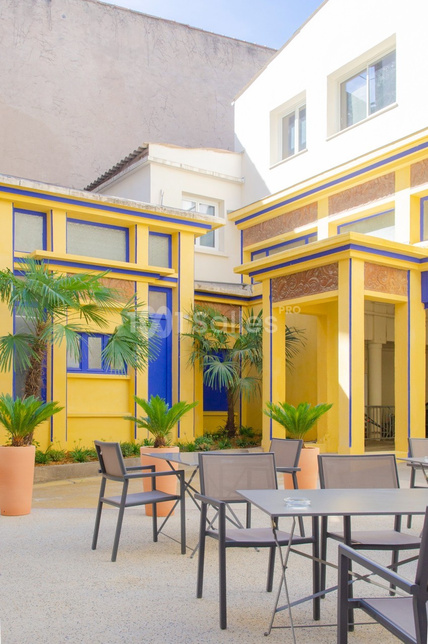 Cour intérieure lumineuse avec façades jaunes et bleues, palmiers en pots et mobilier de terrasse moderne.